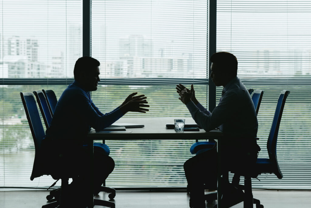 two human outlines against shuttered office window sitting opposite each other negotiating